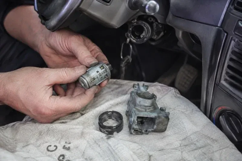 Hands holding a removed ignition cylinder beside a car steering column, with the housing and small parts on a cloth. — Ignition Lock Repair, Vaughan